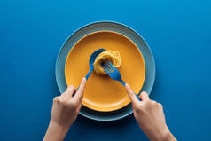 妇女拿着蓝色勺子，在蓝色背景上另一个盘子上方的黄色盘子上用叉子吃的意大利面