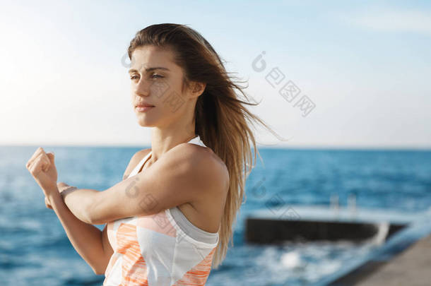 女孩设定目标，保持身体健康，伸展手臂，做慢跑热身练习看起来<strong>坚定</strong>，集中锻炼，站在海边享受码头空气在每天早晨健身跑步