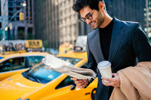 微笑成功的企业家在典雅的西装满意与文章在日报站立在大道与空白咖啡杯, 愉快的商人读报纸等待黄色出租汽车在 Manhatta