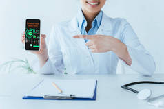 裁剪镜头微笑的拉丁医生用手指指着智能手机与屏幕上的营销分析应用程序