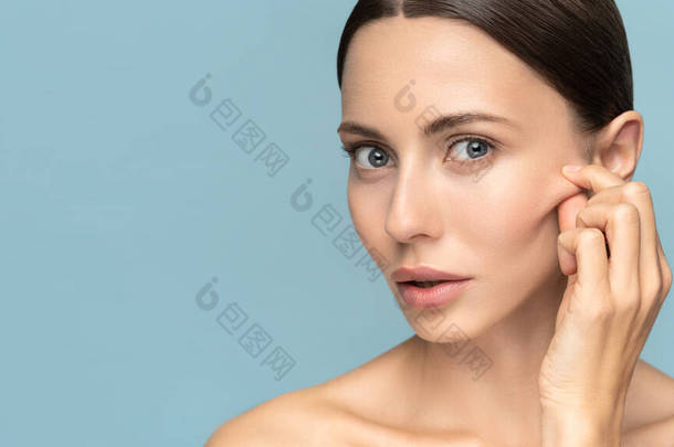 没有化妆的女人在糖醋酸果皮后摸着脸颊，脸上有<strong>老化</strong>的皮肤痕迹，看着相机，在工作室蓝色的背景上被隔离。美容护肤、美容面部治疗