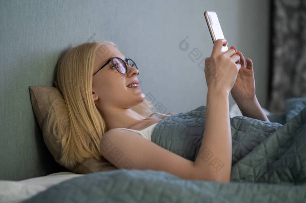 笑着看<strong>手机</strong>的女人躺在白色的床上。快乐的金发年轻女人在家里<strong>用手机</strong>.