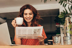 带咖啡的微笑的女人的肖像在桌子上看报纸在咖啡店的笔记本电脑
