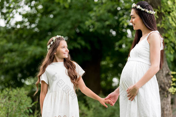 快乐怀孕的母亲和可爱的女儿在白色礼服和花圈牵手和微笑在公园里