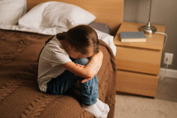 看到一个心烦意乱的小女孩抱着膝盖，低着头哭泣，一个人坐在卧室的床上哭泣的情景，.