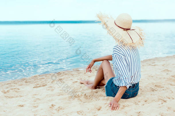在沙滩上放松稻草帽的年轻女子的后视图 