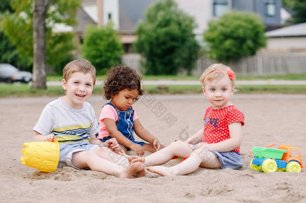 三可爱的白种人和拉美裔儿童的肖像, 孩子们<strong>坐</strong>在沙盒里玩塑料五颜六色的玩具。小<strong>女孩</strong>和男孩朋友一起在操场上玩得开心. 