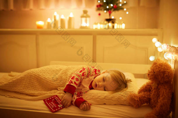 可爱的蹒跚学步的小女孩和泰迪熊睡在圣诞树下，手里拿着给圣诞老人的信。除夕之夜在床上做梦的孩子.