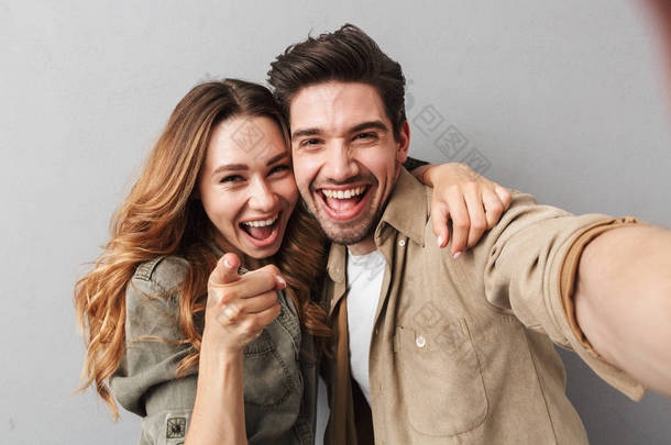 一对快乐的年轻夫妇的肖像拥抱, 而采取了自拍和手指在相机孤立的灰色背景