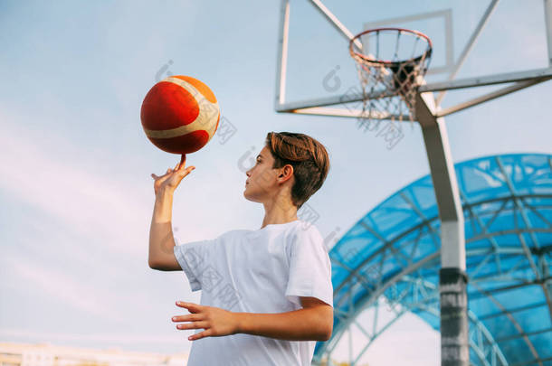 一位身穿白色T恤的少年<strong>篮球</strong>运动员站在<strong>篮球</strong>场上，用手指捻着<strong>篮球</strong>。体育与健康生活方式的概念