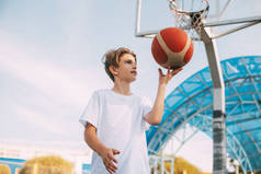 一位身穿白色T恤的少年篮球运动员站在篮球场上，用手指捻着篮球。体育与健康生活方式的概念