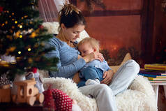 母亲在冬天的圣诞树旁舒适的扶手椅上给幼儿儿子喂奶