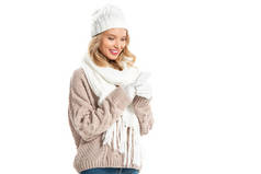 欢快的金发碧眼的女人在冬季毛衣和帽子使用智能手机孤立的白色