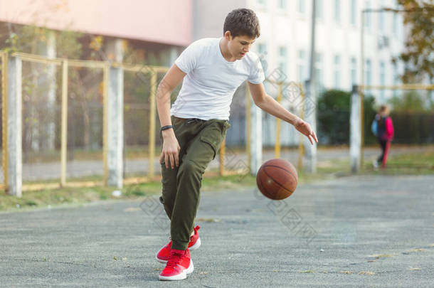 可爱的小男孩在街上的操场上打篮球.穿着白色T恤,外面有橙色篮球运动的青少年.业余爱好、积极的生活方式、儿童体育活动.