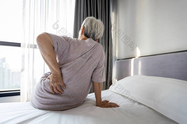 紧张的亚洲老年妇女背痛，坐在床上按摩腰部疼痛，不快乐的老年人睡在不舒服的床垫上或姿势后，早上醒来