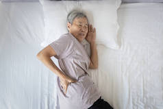 亚洲老年妇女背痛，睡觉时腰酸背痛，不快乐的老年妇女在床上休息时由于姿势不好或床垫不舒服而感到背部肌肉不舒服