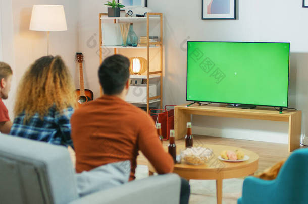 三位朋友坐在沙发上，一边吃小吃一边喝饮料，一边看绿色彩色电视。 在家中<strong>玩乐</strong>的青少年.