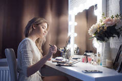神奇的年轻女子在镜子前化妆。化妆品桌旁漂亮姑娘的画像