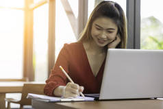 漂亮迷人的亚洲女人，带着手提电脑的微笑，写着成功的想法和要求，在咖啡店、商业概念、暖通等办公室外工作
