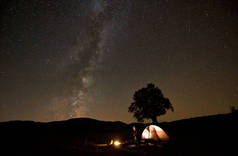 男子坐在旅游帐篷和三脚架相机前燃烧营火, 观看美丽的黑暗星空与银河星座