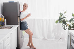 赤脚金发女人站在冰箱边喝茶