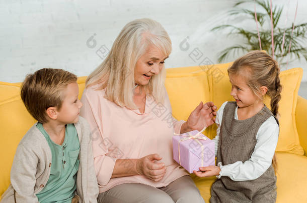 快乐的老妇人和孙子坐在黄色的沙发上，从孙女那里接过礼品盒