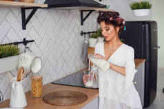 美丽的女孩穿着白色浴袍，在时尚的舒适厨房里，将牛奶从酒杯里倒入玻璃杯中。健康饮食理念.