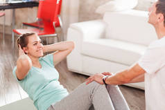 苗条身材的夫妻在家里锻炼得很开心，男人帮助女人在客厅的垫子上做仰卧起坐，丈夫支持妻子锻炼身体，脂肪燃烧腹肌锻炼减肥。横向照片：