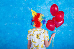 快乐的小丑。假日概念。一个穿着小丑西装的开朗年轻女孩, 在蓝色背景下摆在相机前面。4月1日愚人节的概念。生日.