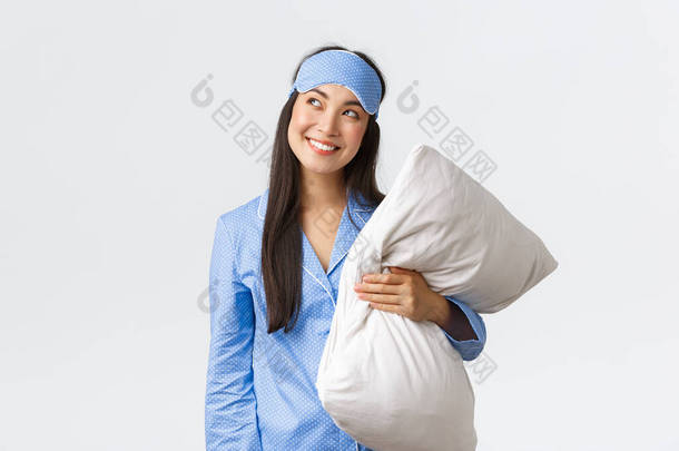 穿着蓝色睡衣，戴着睡眠面罩，头脑发热，心事重重的卡瓦伊亚洲女孩，抱着枕头，看着好奇的左上角，像在想什么，想象什么，白色背景