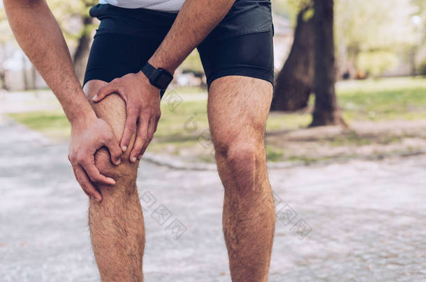 年轻运动员站在公园里和触摸受伤的膝盖的裁剪镜头