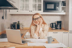 笔记本电脑后面的女人在家里工作。一个穿着夹克和眼镜的金发美女通过视频通话。女学生用爱心、在线培训、梦想工作、自由职业的目光凝视着屏幕.