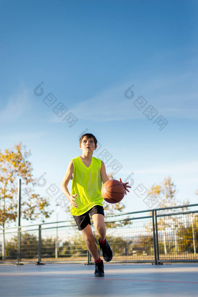 穿着黄色衬衫的可爱男孩在城市操场上打篮球.积极的青少年玩橙色球的户外游戏.