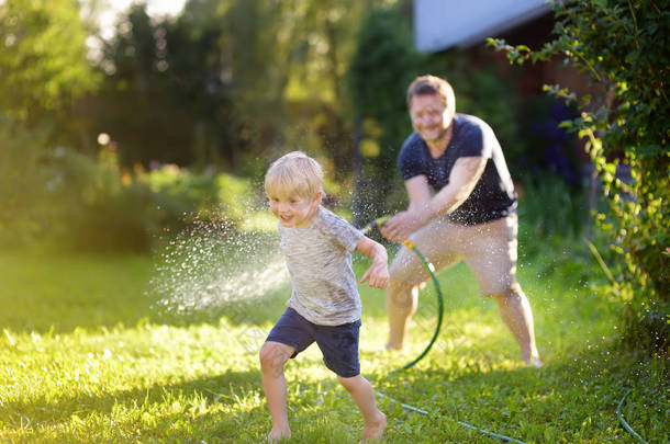 有趣的小男孩与他的父亲玩花园软管在阳光明媚的后院。学龄前儿童在用水喷雾玩乐.