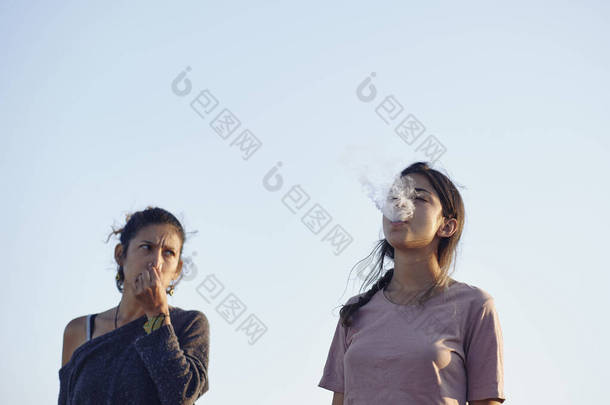 年轻的女人对另一个生气又捂住嘴的女人恼怒地抽烟。一个注意力不集中的女人.