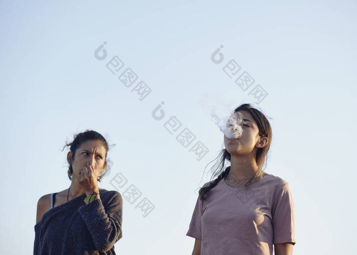 年轻的女人对另一个生气又捂住嘴的女人恼怒地抽烟。一个注意力不集中的女人.