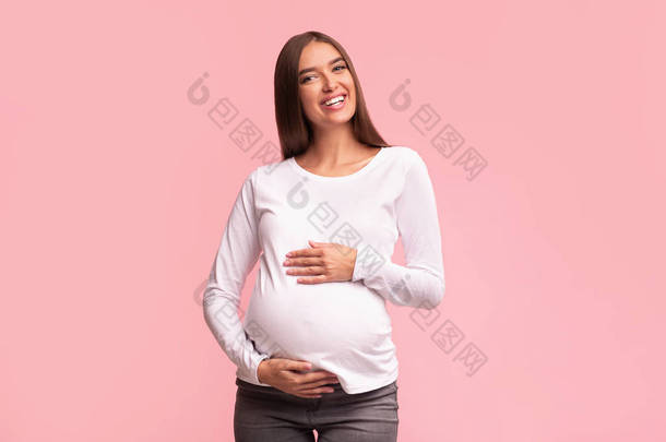 快乐的孕妇触摸着站在粉色背景上的美女
