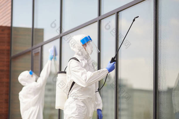 两名穿着<strong>防护服</strong>的工人在建筑物室外消毒或清洁、复制空间时喷洒化学品的肖像
