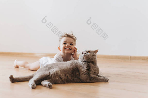 微笑的蹒跚学步的男孩和灰色英国短毛猫猫躺在地板上一起在家