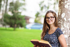 女学生穿着便服和眼镜捧着一本书, 看着你微笑着坐在校园外的绿色草坪树上的背景。多元文化模式, 混合种族, 亚洲俄罗斯女孩