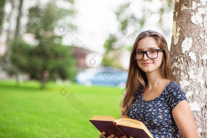 女学生穿着便服和眼镜捧着一本书, 看着你微笑着坐在校园外的绿色草坪树上的背景。多元文化模式, 混合种族, 亚洲俄罗斯女孩