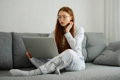 一个戴着眼镜的红头发女孩，穿着睡衣，两条腿交叉坐在沙发上，看着笔记本电脑