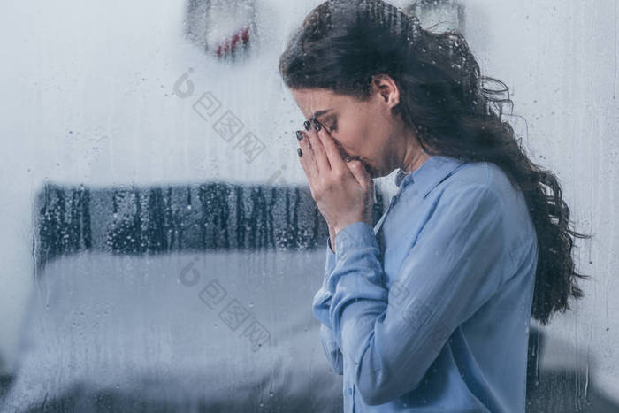 郁闷的女人用双手捂住脸, 用窗户在家里哭着, 用雨滴和复制空间