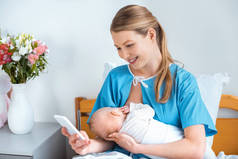 美丽的微笑的年轻母亲母乳喂养婴儿, 并在病房使用智能手机
