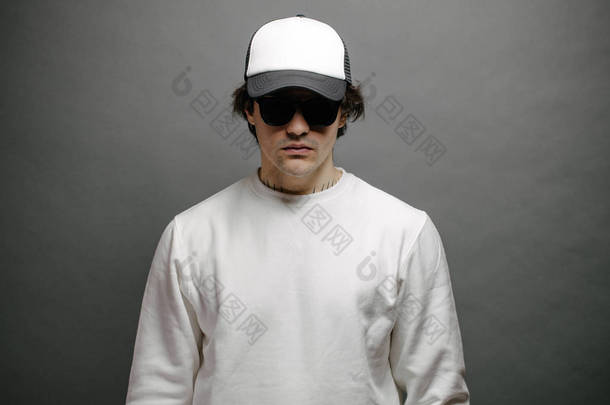 身穿空白白色运动衫，头戴空白棒球帽的男子站在灰色背景之上。运动衫或帽衫，用于模拟、标志设计或有自由空间的设计印刷品.