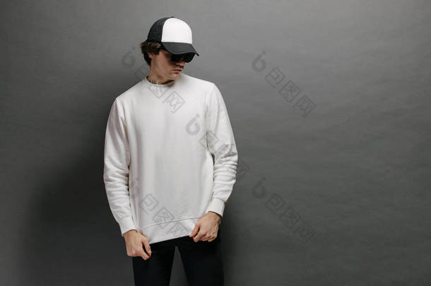身穿空白白色运动衫，头戴空白棒球帽的男子站在灰色背景之上。运动衫或帽衫，用于模拟、标志设计或有自由空间的设计印刷品.