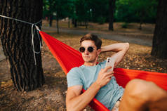 戴着太阳镜、穿着休闲装的英俊而严肃的男人躺在树林里一棵树的吊床上，手里拿着智能手机，满脸专注地看着远方。在公园休息.