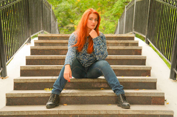 红头发的女孩穿着温暖的秋毛衣坐在城市公园的桥的台阶上