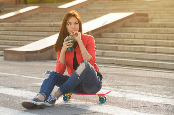 一个少女喝新鲜的水果，坐在滑板上。滑板后的喘息。城市空间的积极生活方式。青年时尚.