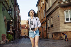 年轻的女摄影师这个女孩的头发是黑色的.在这个古老的欧洲城市，她戴着一顶黑色的帽子，戴着一副太阳镜，带着照相机站在路上.